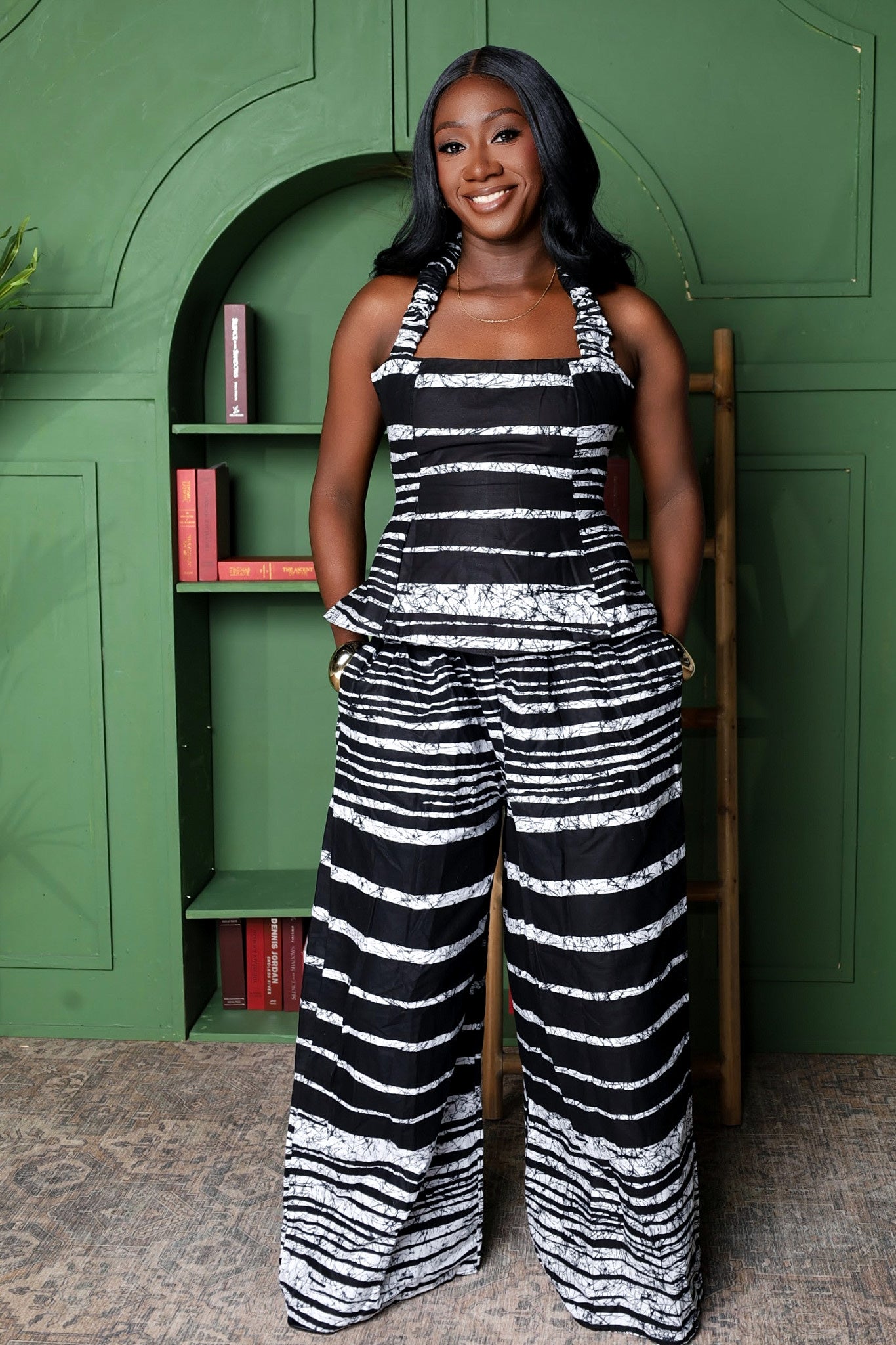 Woman wearing a black and white striped peplum top and wide legged pants standing in front of a green wall with a bookshelf.